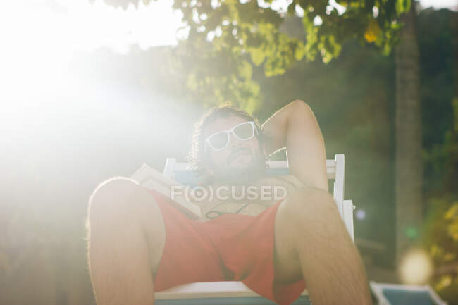 Hombre adulto en gafas de sol descansando en la cama de sol con libro disfrutando de vacaciones de verano en la luz del sol brillante, Tailandia - foto de stock