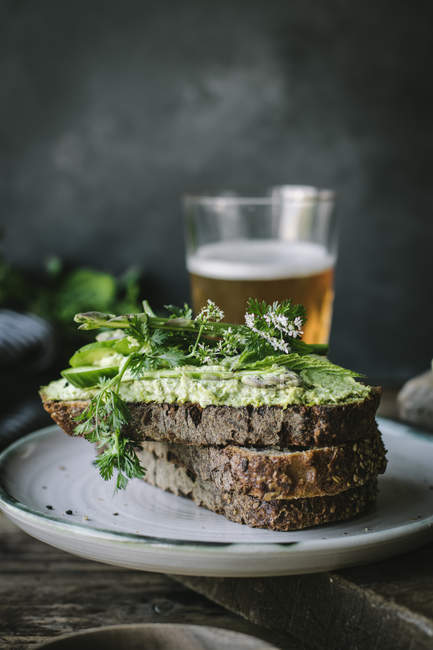 Pane tostato con patè di anacardi verde, erbe e fette di cetriolo con bicchiere di birra su tavola di legno — Foto stock
