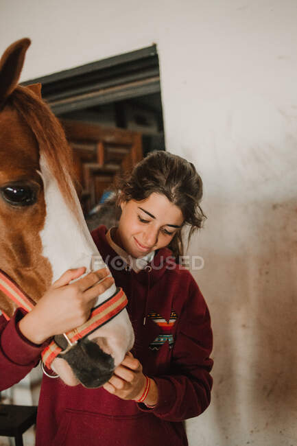 Adolescente chica abrazando con pequeño pony en lindo sombrero en orejas de pie dentro de estable - foto de stock