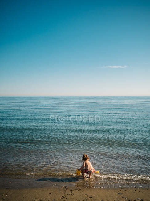 Back view of anonymous cute female toddler playing on sandy shore on background of calm sea — Stock Photo