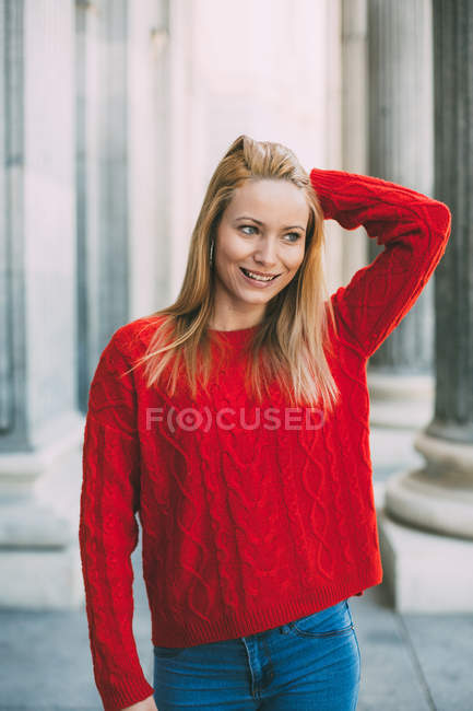 Весела молода жінка в модному червоному светрі, дивлячись далеко, стоячи перед мармуровими колонами на міській вулиці — стокове фото
