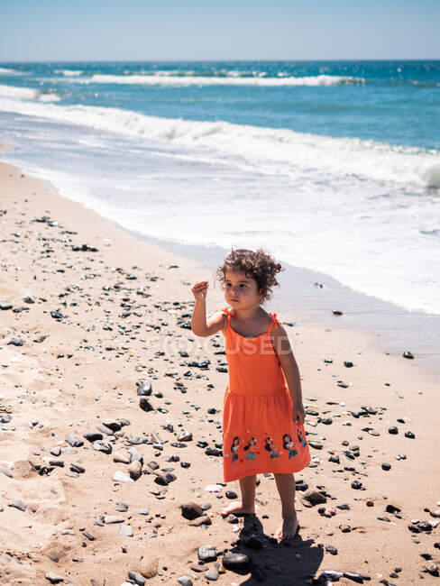 Niña en vestido naranja jugando con una piedra en la playa de arena y mirando el mar y el cielo - foto de stock
