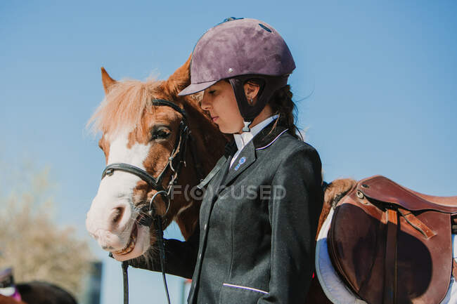 Vista lateral de joven adolescente mujer en casco de jinete y chaqueta acariciando caballo de pie juntos al aire libre contra el cielo azul - foto de stock