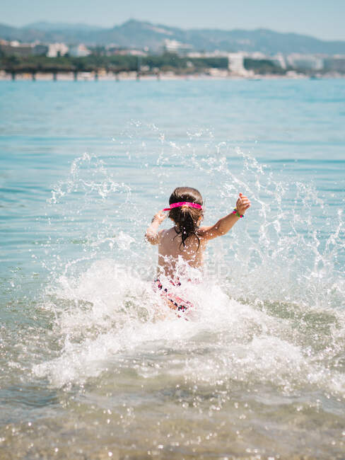 Vue arrière de fille mignonne joyeuse anonyme jouant dans l'eau de mer sur fond de bord de mer calme — Photo de stock