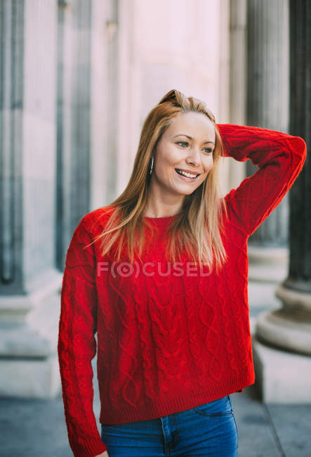 Веселая молодая женщина в модном красном свитере смотрит в сторону, стоя перед мраморными колоннами на городской улице — стоковое фото
