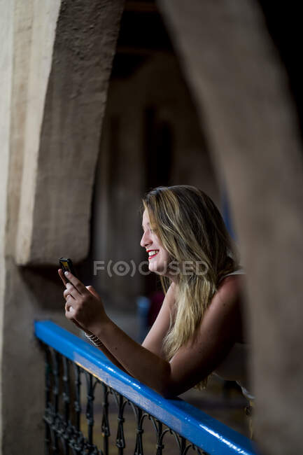 Весела красива жінка в стильному одязі посміхається і переглядає смартфон, спираючись на балконні поручні стародавнього будинку в Марокко. — стокове фото