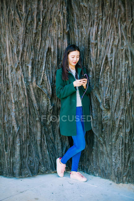 Красивая азиатка в стильном наряде слушает музыку и использует смартфон, опираясь на грубую стену с рельефом деревьев — стоковое фото