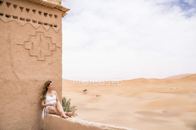 Hermosa joven en la parte superior blanca sentada en la valla de piedra en el viento mirando hacia otro lado contra el desierto de arena sin fin, Marruecos - foto de stock