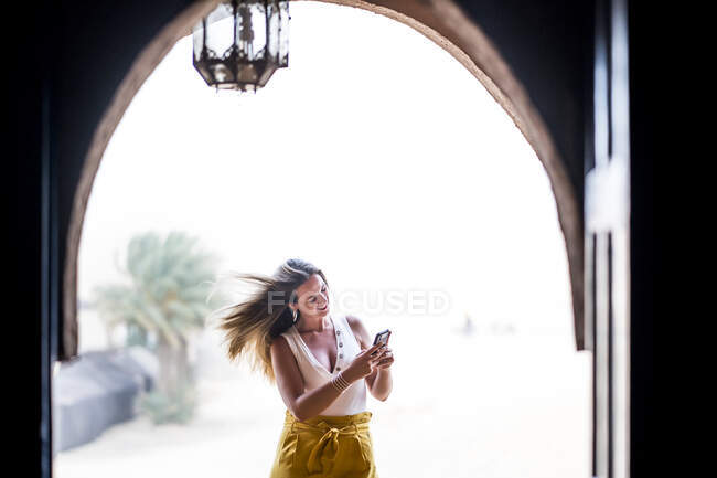 Donna felice utilizzando il telefono cellulare vicino paesaggio desertico in piedi sul balcone di pietra, Marocco — Foto stock