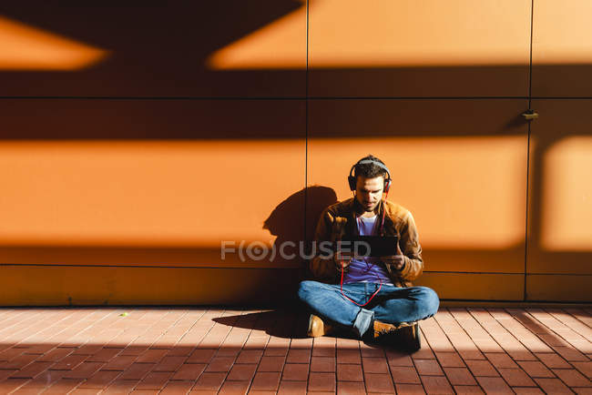 Красивый человек в наушниках слушает музыку и просматривает планшет, сидя на лестнице возле современного здания в солнечный день — стоковое фото