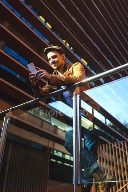 Positiver Mann in stylischem Outfit mit Handy während er an einem sonnigen Tag im modernen Glasbalkon auf einem modernen Gebäude steht — Stockfoto