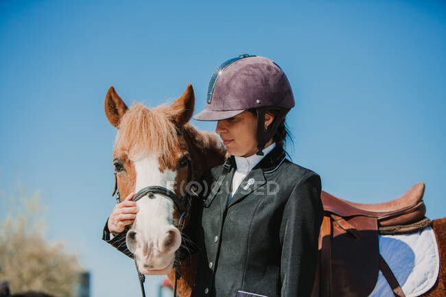 Вид сбоку на молодую девушку-подростка в шлеме жокея и ласкающую лошадь, стоящую на открытом воздухе под голубым небом — стоковое фото