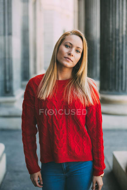 Mujer joven en suéter rojo de moda mirando a la cámara mientras está de pie frente a columnas de mármol en la calle de la ciudad - foto de stock