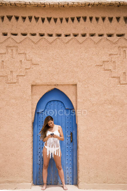 Joyeux jeune femme portant un haut blanc avec bikini et utilisant le téléphone contre la porte orientale bleue dans un mur de pierre, Maroc — Photo de stock