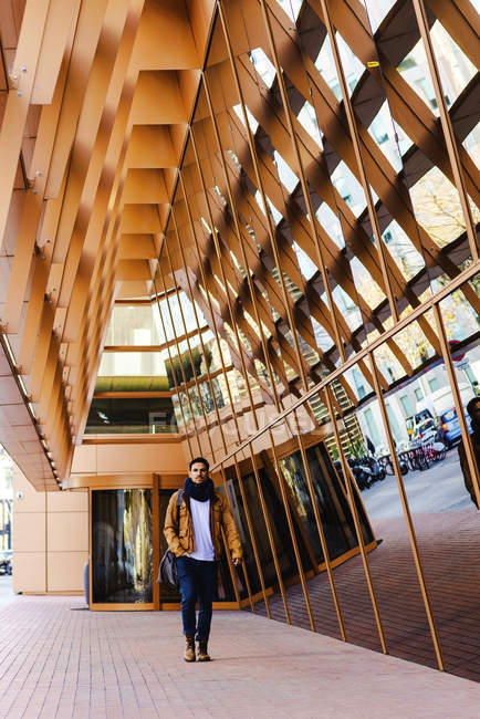 Homme à la mode en tenue décontractée marchant près du mur de verre du bâtiment contemporain — Photo de stock