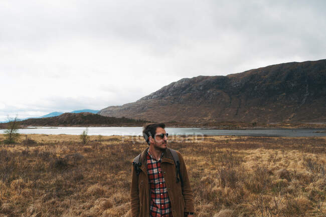 Homem adulto com mochila em pé no pitoresco vale remoto com montanhas e lago olhando para longe — Fotografia de Stock