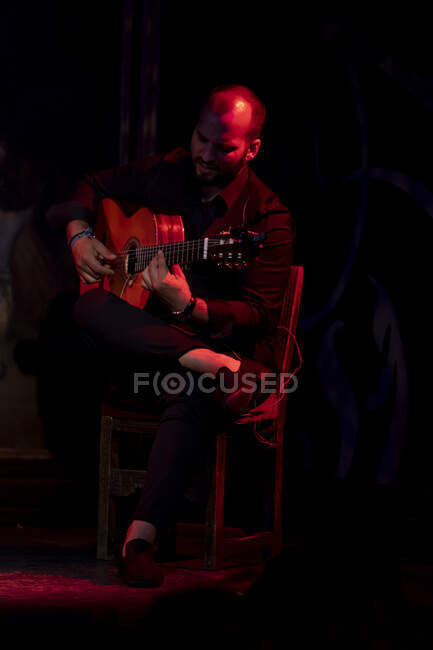 Лысый бородатый парень играет на акустической гитаре, сидя на сцене под красным светом во время выступления фламенко — стоковое фото