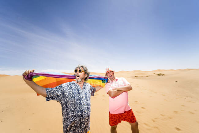 Heureux les hommes mûrs en surpoids qui s'amusent avec le drapeau LGBT tout en se tenant sur fond de désert aride et de ciel bleu — Photo de stock
