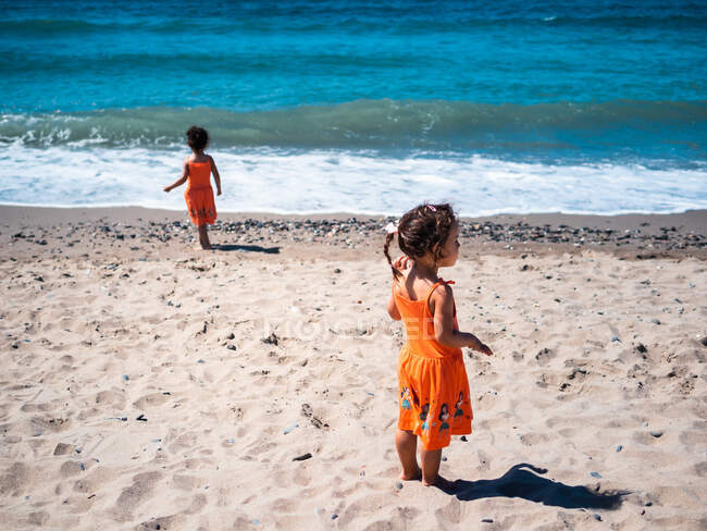 Female kids in orange dress standing on sandy seaside and looking at sea and sky — Stock Photo