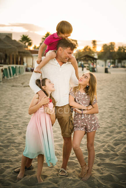 Взрослый мужчина со смеющимся мальчиком на плечах, стоящий с красивыми маленькими девочками на пляже и проводящий время вместе — стоковое фото