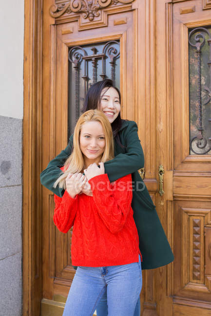 Mujer asiática feliz sonriendo y mirando hacia otro lado mientras está de pie contra la puerta ornamental del edificio envejecido y abrazando amigo caucásico - foto de stock