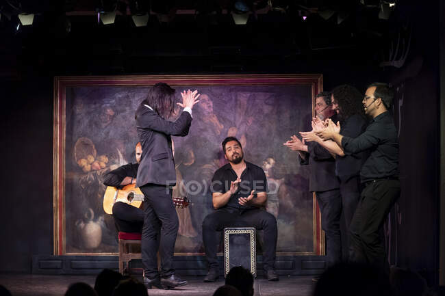 Mann in schwarzem Kostüm tanzt Flamenco in der Nähe von hispanischen männlichen Musikern während einer Performance gegen Malerei auf dunkler Bühne — Stockfoto