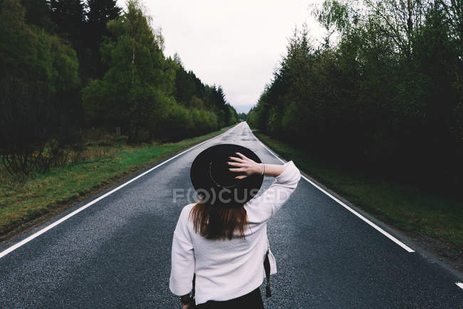 Vue arrière de la femme branchée en chapeau noir debout dans la solitude sur une route reculée avec des arbres verts luxuriants, Écosse — Photo de stock