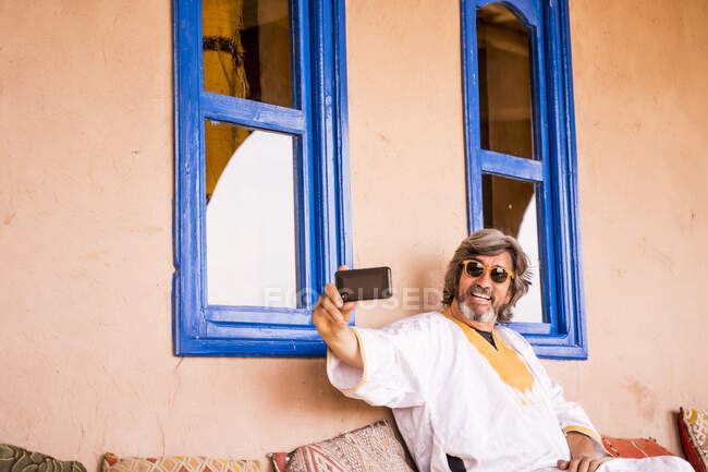 Hombre adulto alegre en ropa larga sentado en el sofá y el uso de teléfono en la casa decorada en estilo oriental, Marruecos - foto de stock