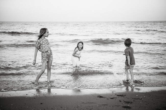 Group of little boy with two sisters playing in shallow wave of water on coast, black and white photo — Stock Photo