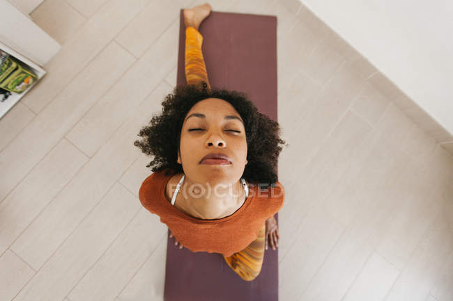 Rosto da jovem mulher afro-americana sentada em pose de ioga com olhos fechados no tapete na sala de luz — Fotografia de Stock