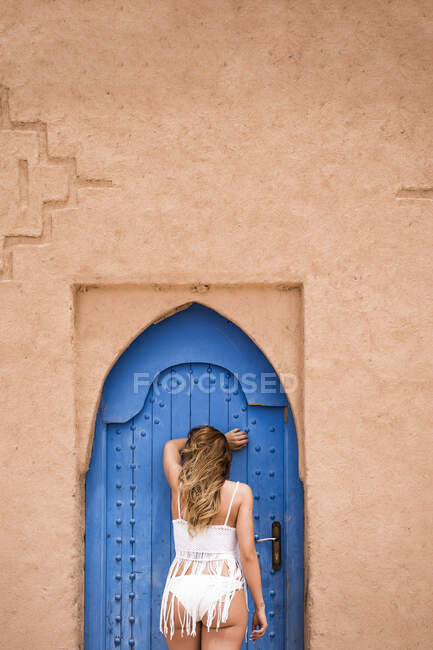 Vista posteriore della donna anonima che indossa top bianco con bikini appoggiato alla porta orientale blu in muro di pietra, Marocco — Foto stock