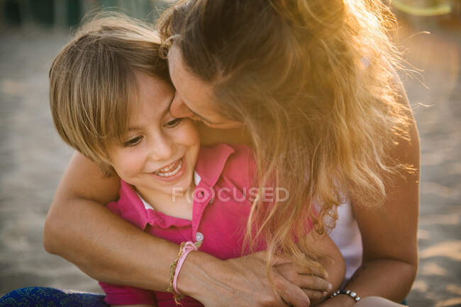 Mother embracing and kissing cute cheerful boy while sitting together on beach in bright sunshine — Stock Photo