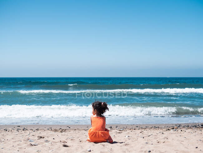 Vista trasera del niño femenino en vestido naranja sentado en la playa de arena y mirando al mar y el cielo - foto de stock