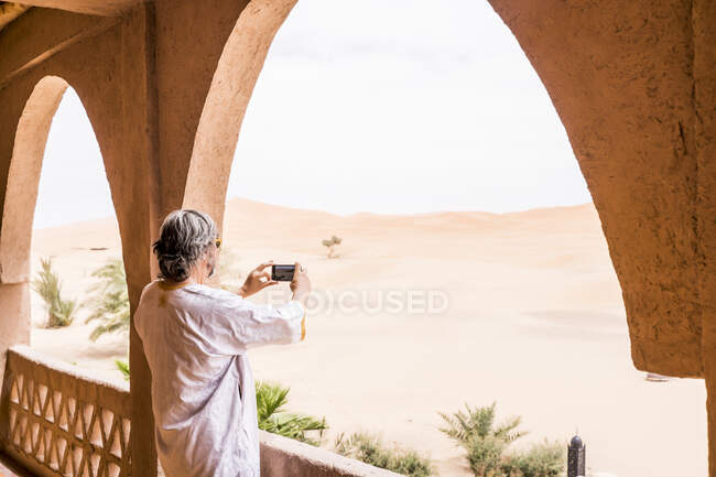 Vista lateral del hombre adulto en traje largo que representa el paisaje del desierto de pie en el balcón de piedra, Marruecos - foto de stock