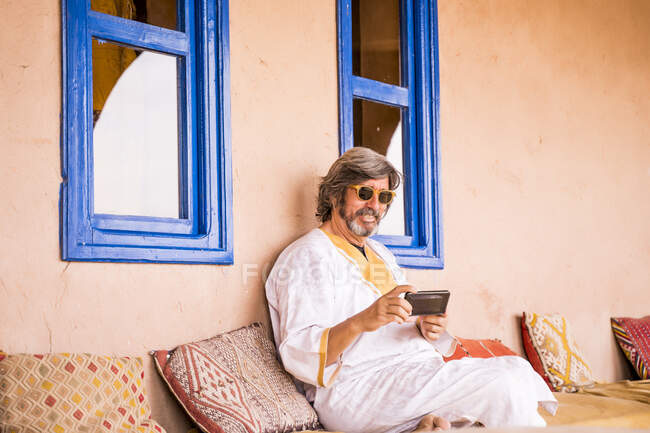 Uomo adulto in abiti lunghi seduto sul divano e utilizzando il telefono a casa decorata in stile orientale, Marocco — Foto stock
