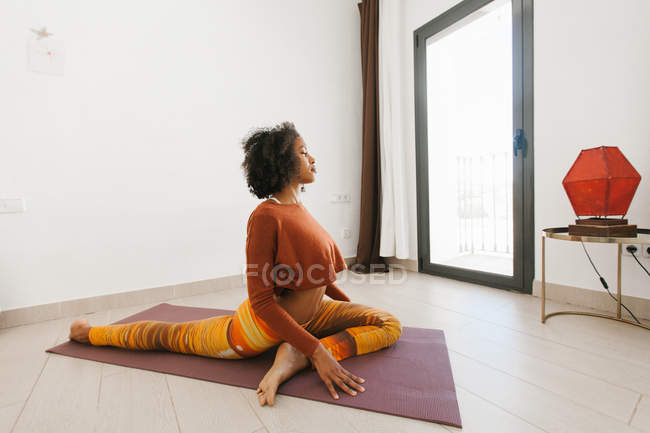 Mujer joven atractiva afroamericana sentada en postura de yoga con los ojos cerrados en la estera en la sala de luz - foto de stock