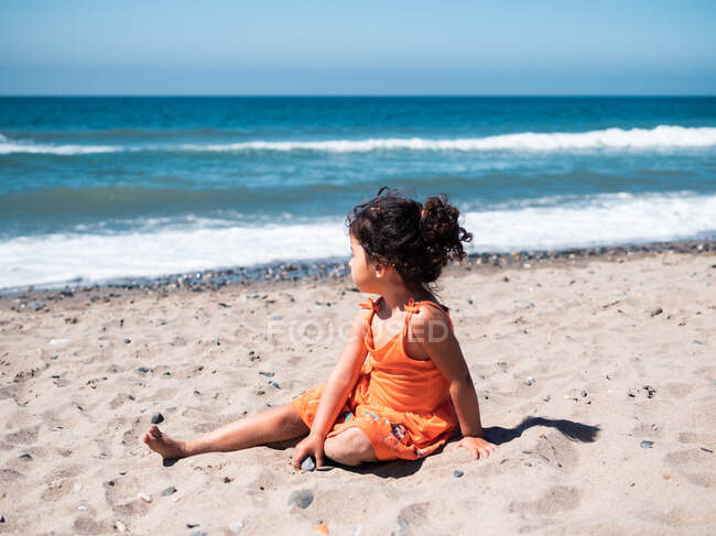 Vista posteriore del bambino femmina in abito arancione seduto sul mare sabbioso — Foto stock