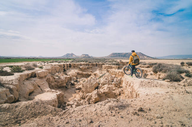 Side view of young man in yellow jacket and backpack with bicycle standing on stony hill stretchering high between stony hills in semi-desert Bardenas Reales Navarra Spain — Stock Photo