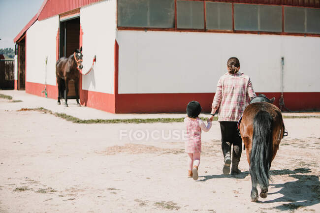 Rückansicht einer Frau, die kleines Mädchen und Pony auf dem Hof des Hippodroms im hellen Sonnenlicht führt — Stockfoto