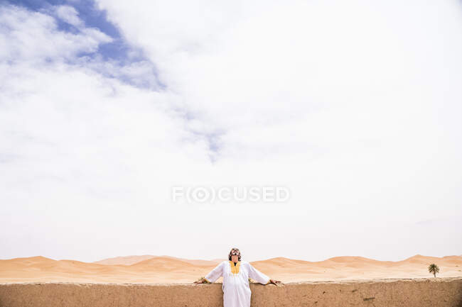 Homme adulte joyeux reposant sur une clôture en pierre sur la terrasse contre un désert sans fin, Maroc — Photo de stock