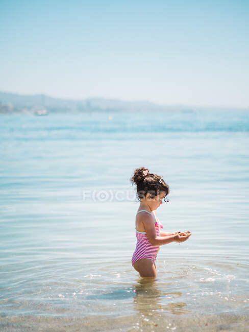 Вид збоку чарівна дівчинка-малюк у купальнику, що стоїть у теплій воді спокійного моря — стокове фото