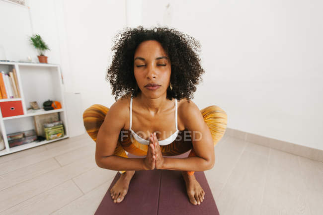 Mujer joven afroamericana realizando pose de yoga con los ojos cerrados agachados sobre una alfombra en casa - foto de stock