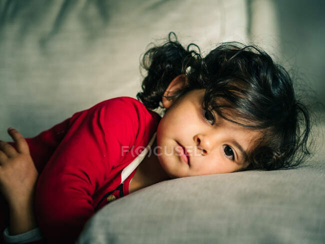 Adorable niña en vestido rojo sonriendo mientras descansa en la almohada en casa - foto de stock