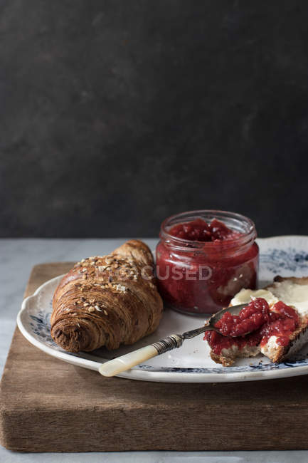 Crujiente croissant y tostadas con mantequilla y mermelada de fresa servidas en plato sobre tabla de madera - foto de stock
