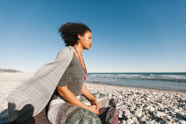 African American young woman meditating in lotus yoga posture on sandy beach in bright day — Stock Photo