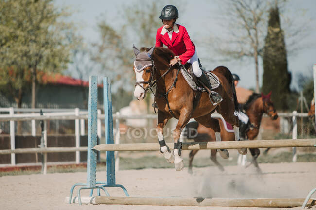 Teenage jockey on horse leaping over horizontal wooden bars while riding on racetrack — Stock Photo