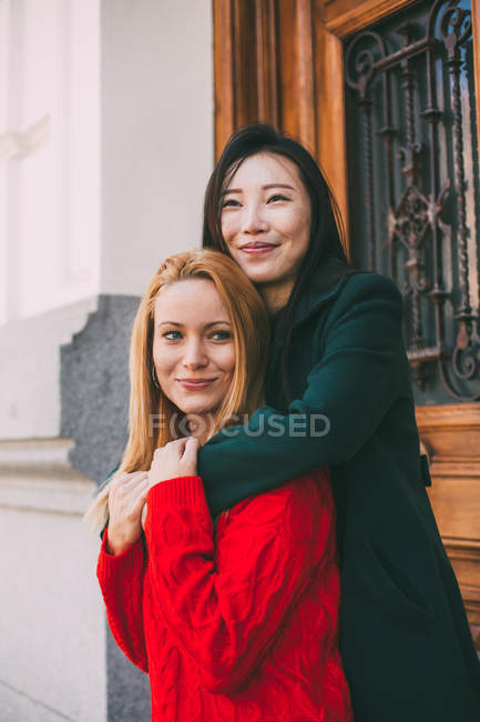 Happy Asian female smiling and looking away while standing against ornamental door of aged building and embracing Caucasian friend — Stock Photo