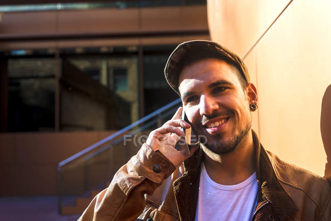 Uomo positivo in abito elegante parlare sul telefono cellulare mentre appoggiato sulla parete dell'edificio moderno nella giornata di sole — Foto stock