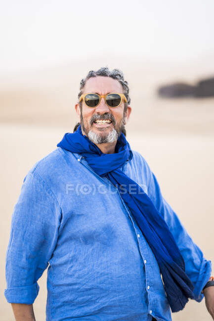 Зрелый бородатый мужчина смотрит в камеру во время прогулки по песчаной пустыне против серого неба в Марокко — стоковое фото