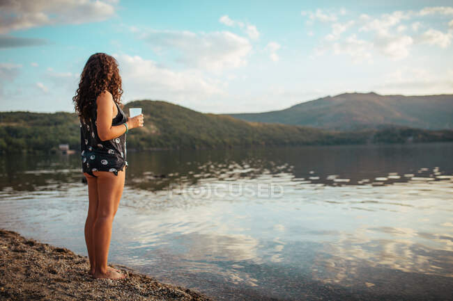 Seitenansicht einer unkenntlichen Frau, die auf einem Felsen im Wasser steht und die Berge im Sonnenuntergang betrachtet — Stockfoto